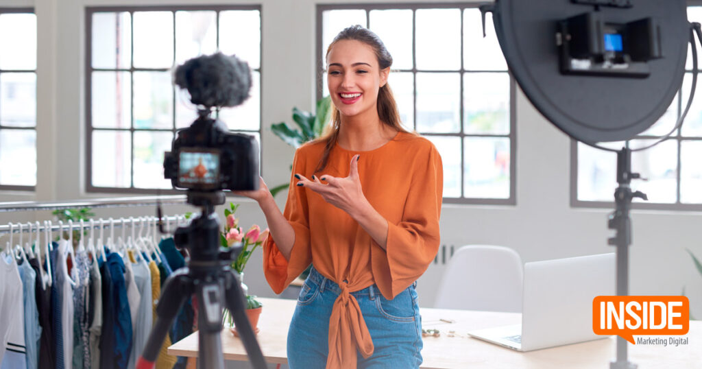 Una mujer está parada frente a una cámara y un aro de luz, hablando y haciendo gestos con una mano. Está en una habitación con un perchero, una mesa con flores y un ordenador portátil. Se ve el texto "INSIDE Marketing Digital".