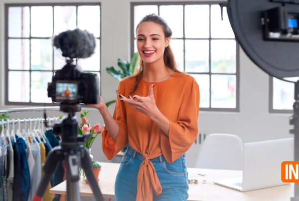 Una mujer está parada frente a una cámara y un aro de luz, hablando y haciendo gestos con una mano. Está en una habitación con un perchero, una mesa con flores y un ordenador portátil. Se ve el texto "INSIDE Marketing Digital".