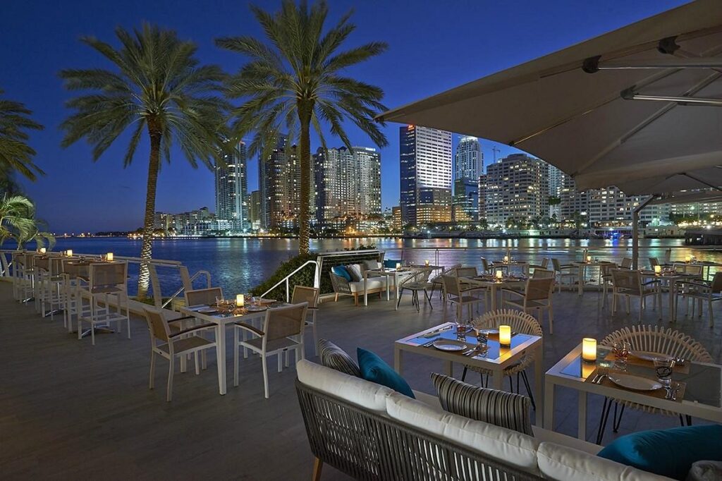 Restaurante al aire libre con palmeras a lo largo de la costa. Las mesas están dispuestas con velas y el horizonte de la ciudad se puede ver al fondo bajo el cielo nocturno.