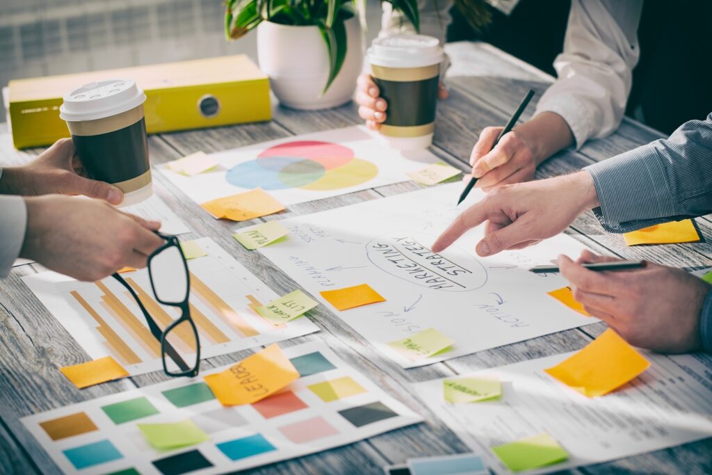 Un grupo de personas trabaja en una mesa de madera cubierta con materiales de marketing, gráficos y notas adhesivas mientras sostienen tazas de café y escriben notas.