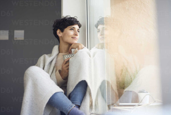 Una persona de cabello corto sostiene una taza envuelta en una manta y mira por la ventana con expresión reflexiva, contemplando el poder del sonido para la relajación y el bienestar.