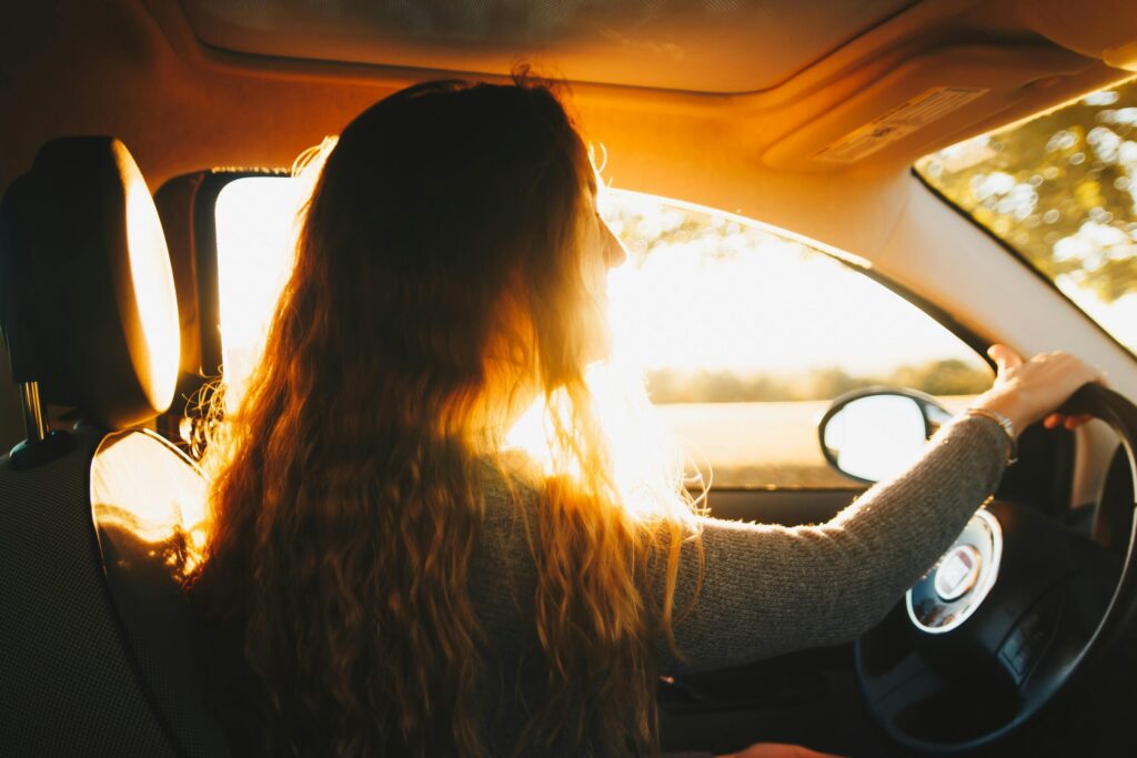 Una persona con el pelo largo y ondulado conduce un coche al atardecer. La luz del sol entra por la ventanilla del coche e ilumina su pelo.