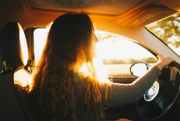 Una persona con el pelo largo y ondulado conduce un coche al atardecer. La luz del sol entra por la ventanilla del coche e ilumina su pelo.