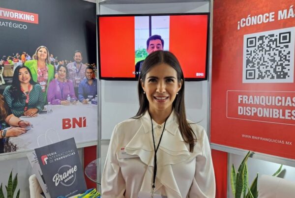 Una mujer con un cordón sonríe frente a un stand. El stand cuenta con carteles con texto sobre redes y franquicias. También hay un código QR y una pantalla de televisión que muestra a una persona.