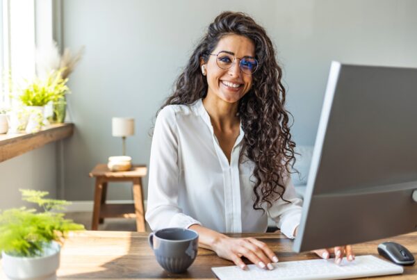 Una mujer con cabello largo y rizado y anteojos sonríe mientras trabaja con una computadora. Está sentada en un escritorio de madera con una taza de café a su lado en una habitación bien iluminada con plantas.