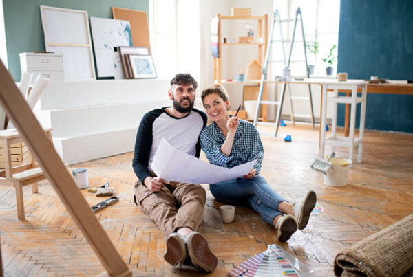 Una pareja se sienta en el suelo de una habitación en proceso de renovación, revisando una gran hoja de papel que detalla su plan de mejoras, rodeada de materiales y herramientas para pintar. Planificando cuidadosamente la estrategia, miran de vez en cuando el documento que describe los detalles de sus préstamos para reforma de vivienda.