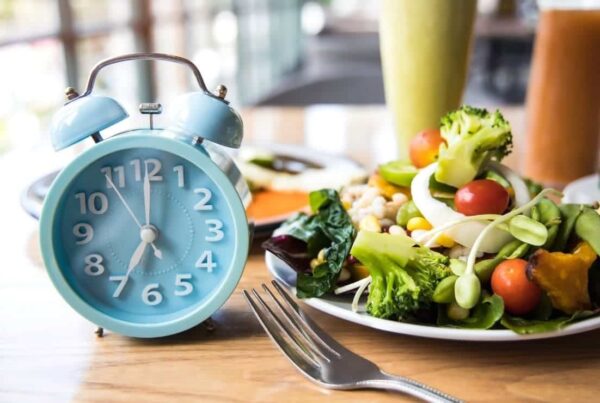 Un reloj despertador azul se encuentra junto a un plato de ensalada fresca con tomates, brócoli y verduras en una mesa de madera, junto con un tenedor.