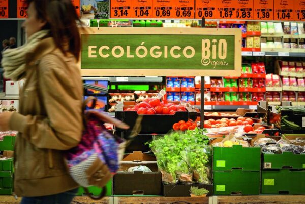 Una persona pasa junto a un escaparate de un supermercado con la etiqueta "Ecológico Bio Organic" con diversas frutas y verduras, incluidos tomates, rábanos y verduras de hoja verde.