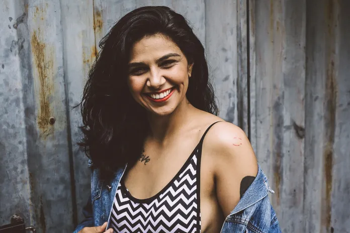 Mujer con cabello largo y oscuro sonriendo, vistiendo una blusa de chevron blanco y negro y una chaqueta de mezclilla, parada frente a una pared de metal corrugado.