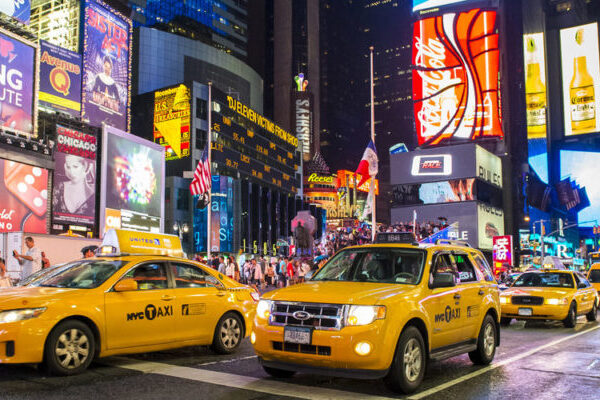 Times Square iluminada por la noche con numerosos taxis amarillos y anuncios luminosos. Se ven peatones en las aceras.