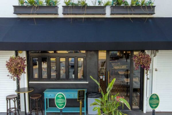 Vista de la calle de una tienda de sándwiches llamada "Sanguich" con un toldo negro, asientos al aire libre y plantas decorativas.