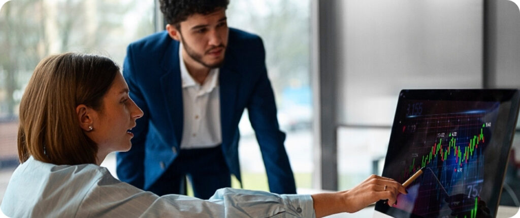 Dos personas en una oficina miran una pantalla de computadora que muestra un gráfico financiero con barras y líneas ascendentes. Una persona señala la pantalla con un lápiz.