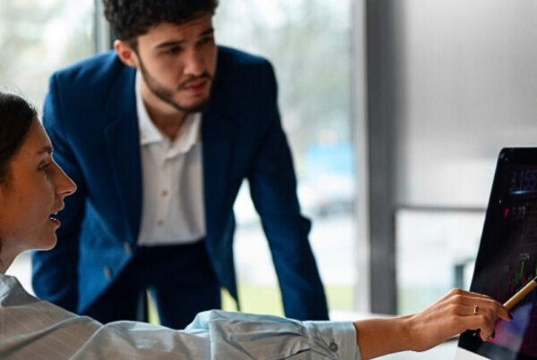 Dos personas en una oficina miran una pantalla de computadora que muestra un gráfico financiero con barras y líneas ascendentes. Una persona señala la pantalla con un lápiz.