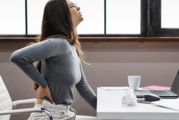 Una mujer sentada en un escritorio sostiene su espalda baja con incomodidad, sentada cerca de una computadora portátil y una taza.