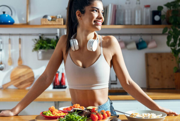 Una mujer sonríe en una cocina, con auriculares colgados del cuello. Está de pie detrás de una mesa con platos de frutas y verduras frescas.