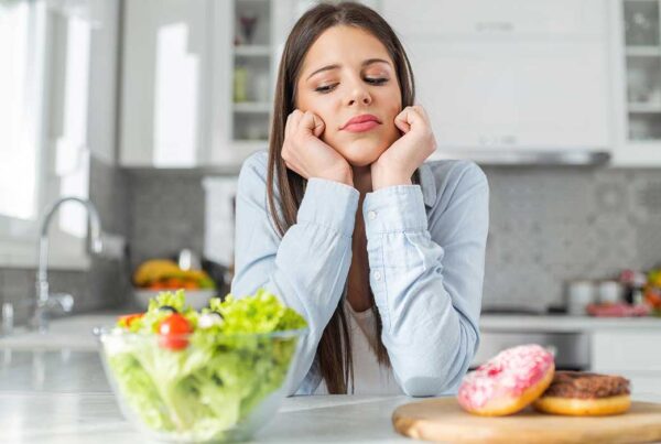 Una mujer está sentada en el mostrador de la cocina, mirando una ensaladera y dos donas con expresión pensativa.